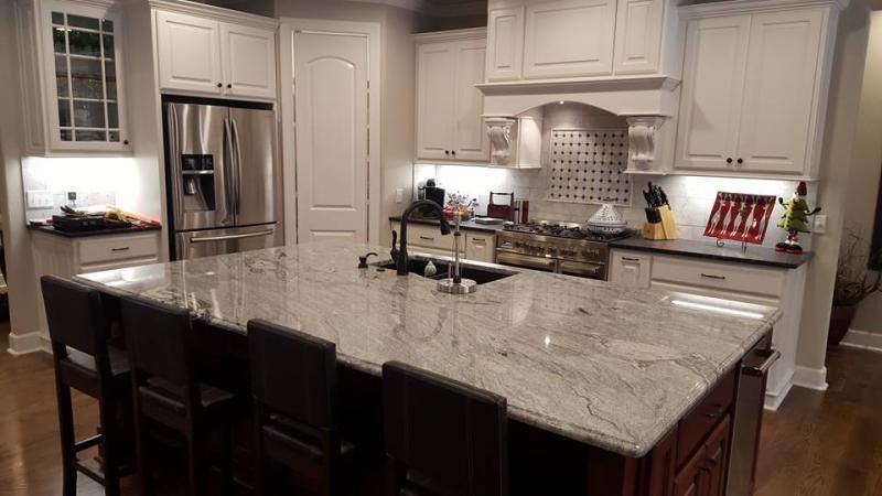 A spacious kitchen with white cabinets, a large granite island with bar stools, stainless appliances, and wood flooring.