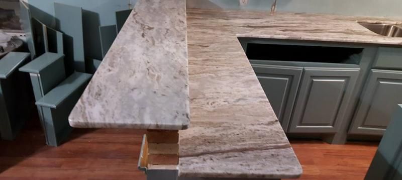 An L-shaped granite countertop with a raised bar section, set above sage green kitchen cabinets and wooden flooring.
