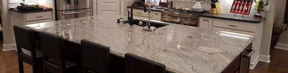 A large kitchen with a dark-base island topped with light gray, veined granite and several dark chairs.