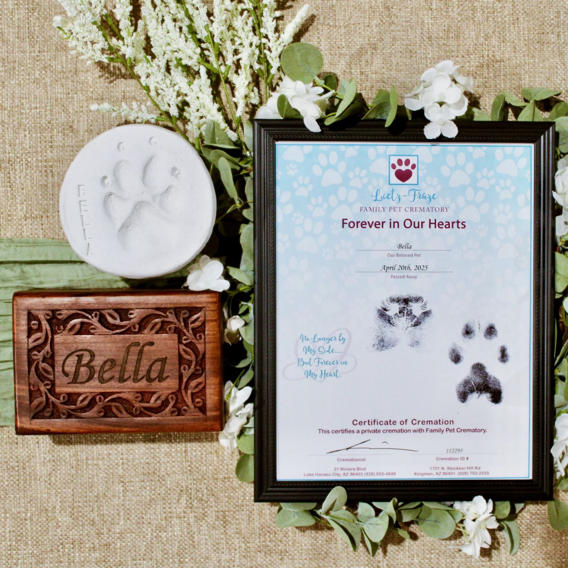 Memorial plaque with paw prints beside a carved wooden box labeled Bella, surrounded by white flowers