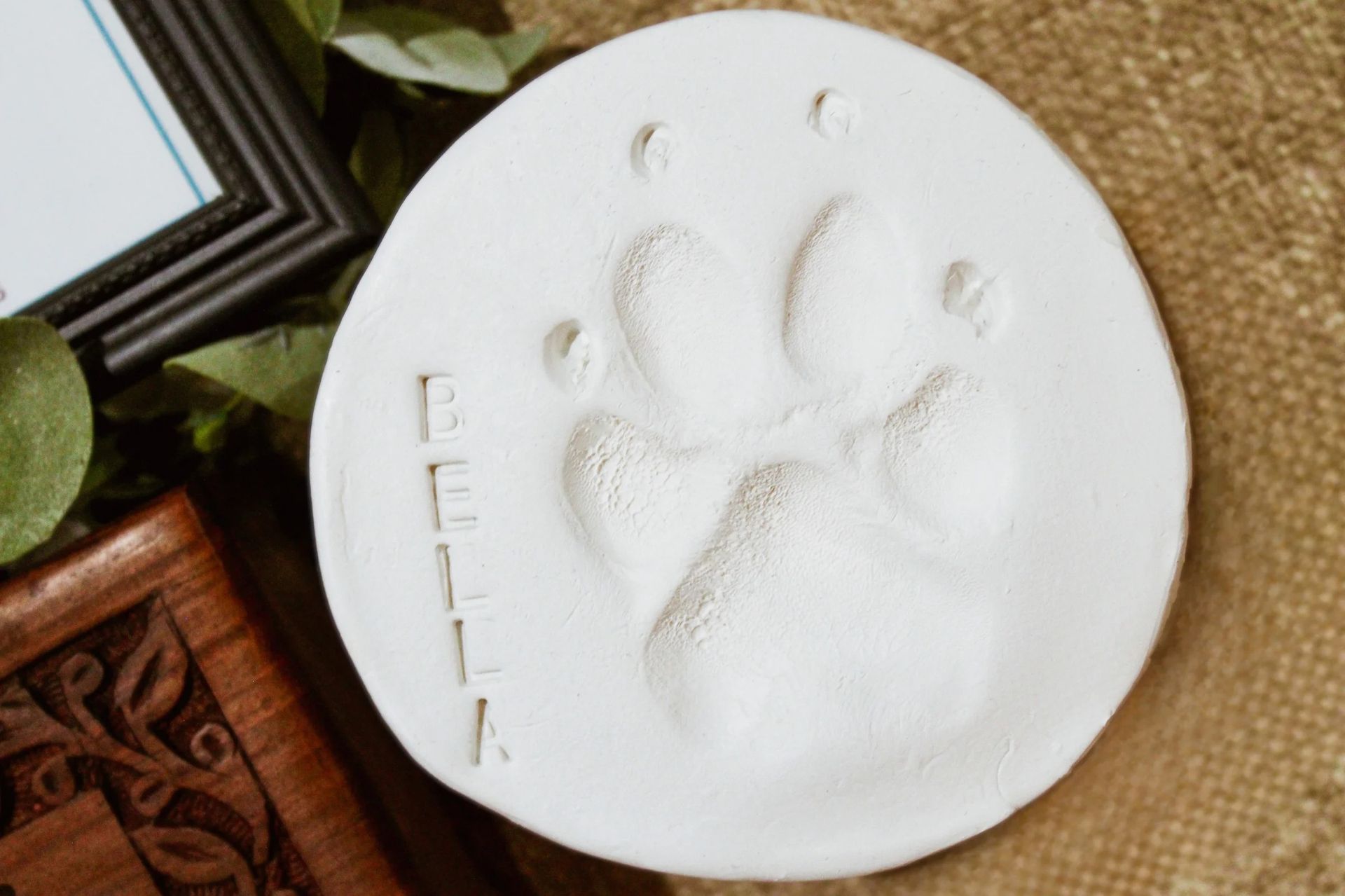 White ceramic paw-print plaque with “BELLA” engraved, resting on a woven mat.