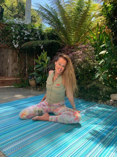Woman seated cross-legged on a blue striped mat in a sunny garden, smiling amid lush green plants.