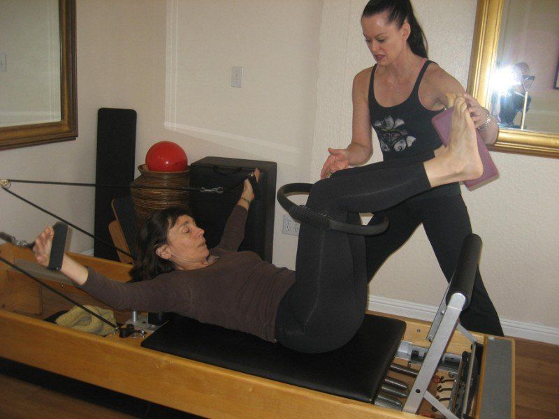 Two people exercising on a Pilates reformer in a studio, one assisting the other.