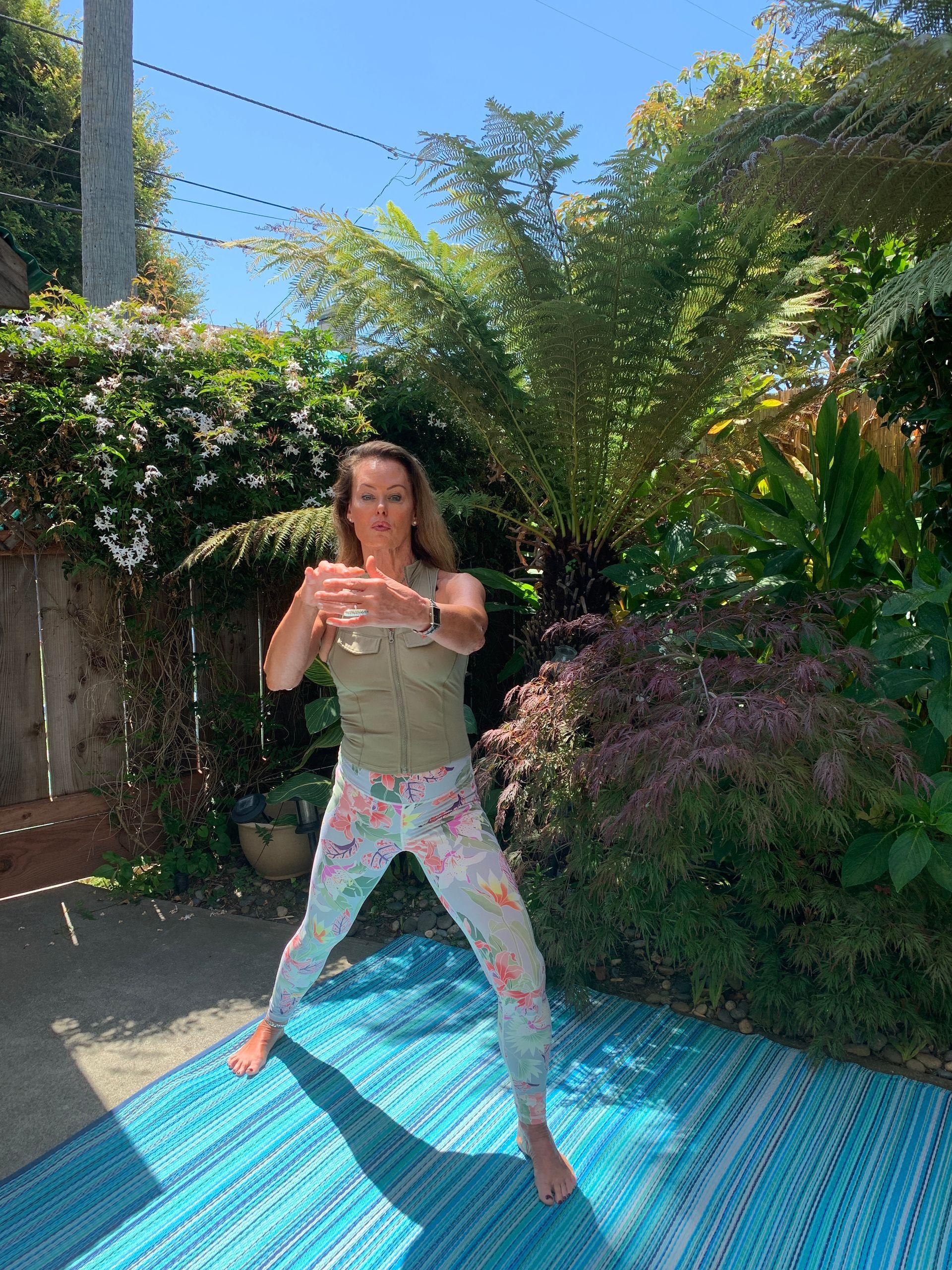 Person stretching on a blue outdoor mat in a sunny garden with lush greenery and flowers