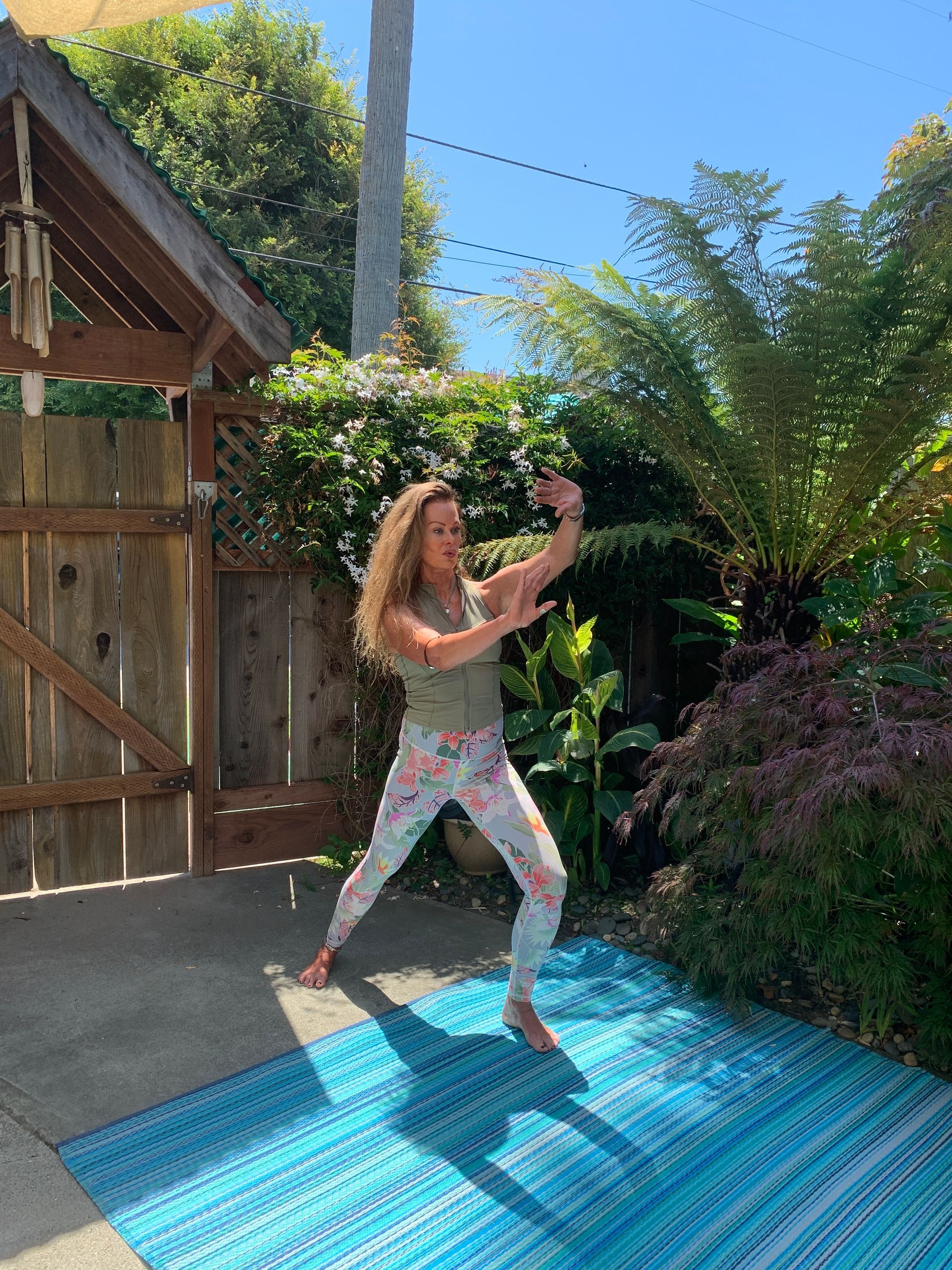Woman posing on a blue rug in a sunny backyard garden, near a wooden gate and greenery.