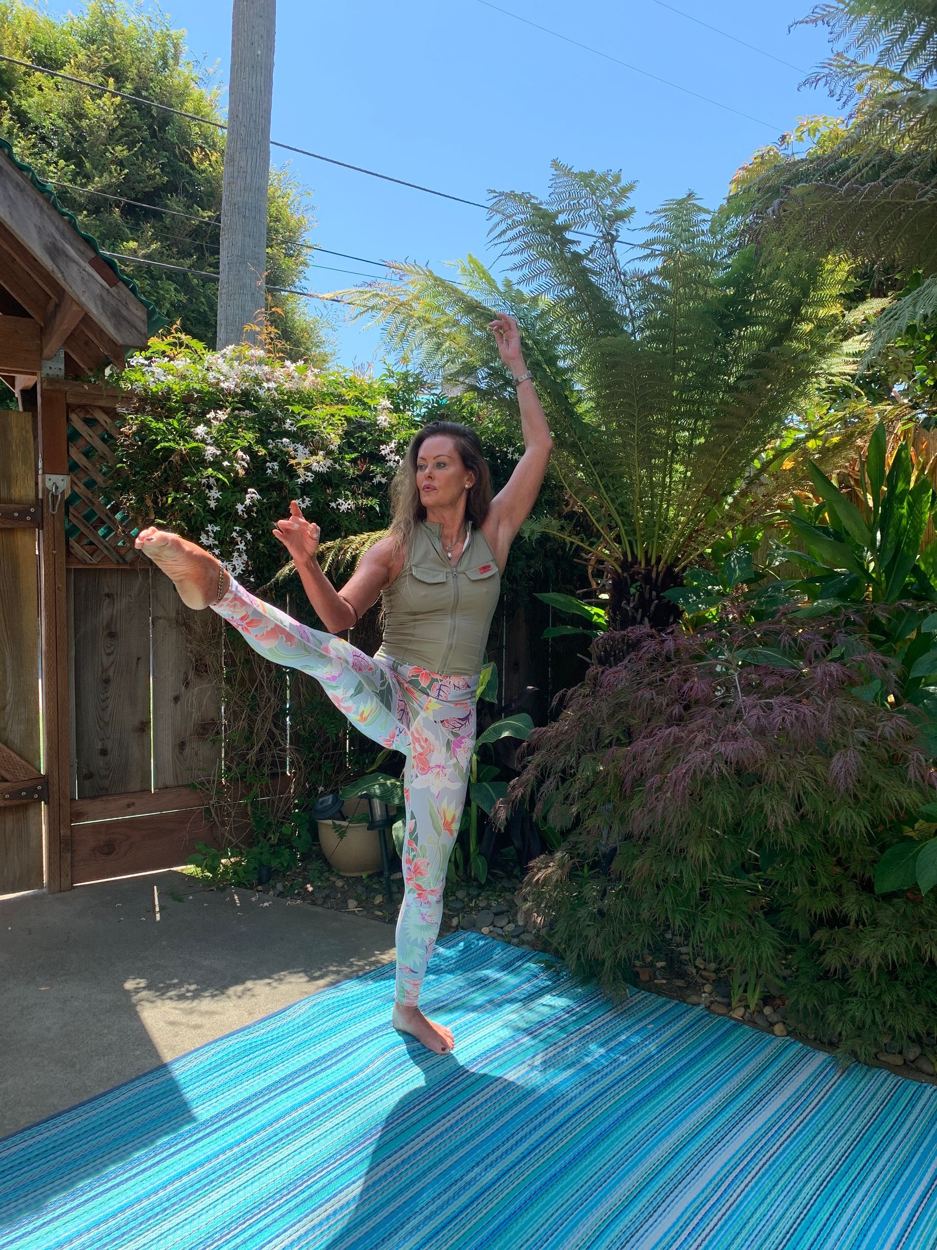 Person balancing in a high leg pose on a blue mat in a sunny garden courtyard