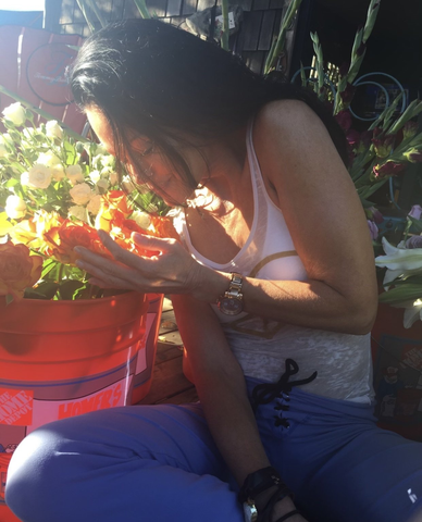 Person smelling flowers in a red bucket in bright sunlight, sitting among potted plants.