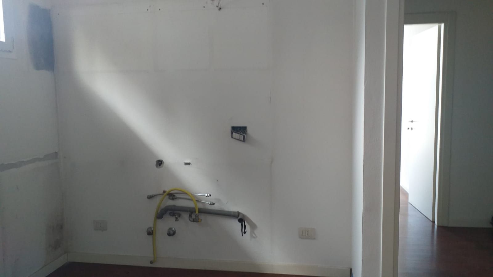 Stanza vuota con pareti bianche, tubature a vista e una porta aperta.