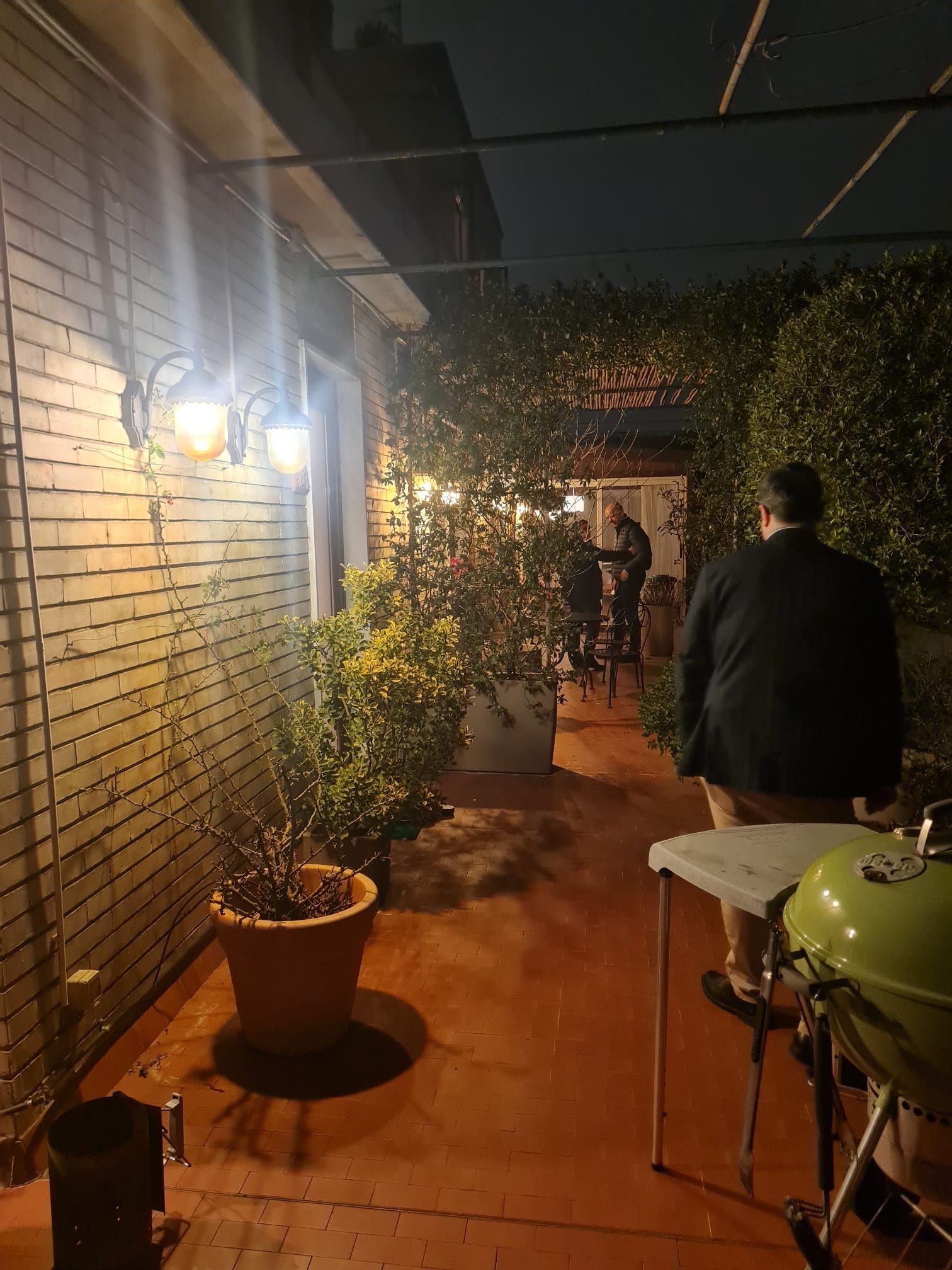 Patio in mattoni con lucine, piante e griglia. Due persone sono in piedi vicino a un tavolo in fondo al vialetto.