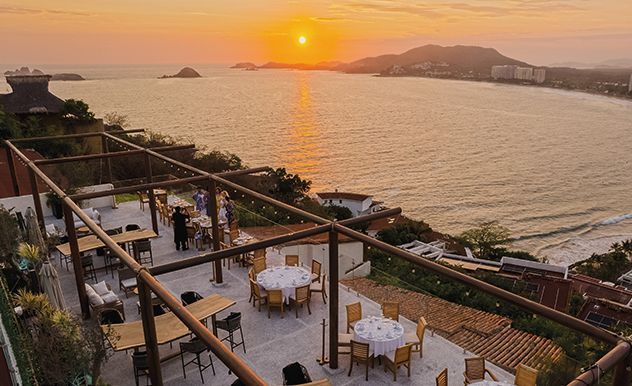 Puesta de sol sobre una terraza junto al mar con mesas, gente y vistas al océano, bañada por una cálida luz anaranjada.
