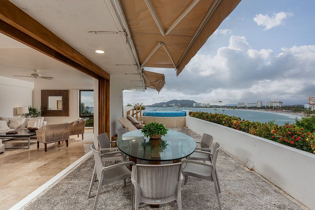 Patio cubierto junto al mar con mesa de cristal, sillas y vistas al océano al atardecer.