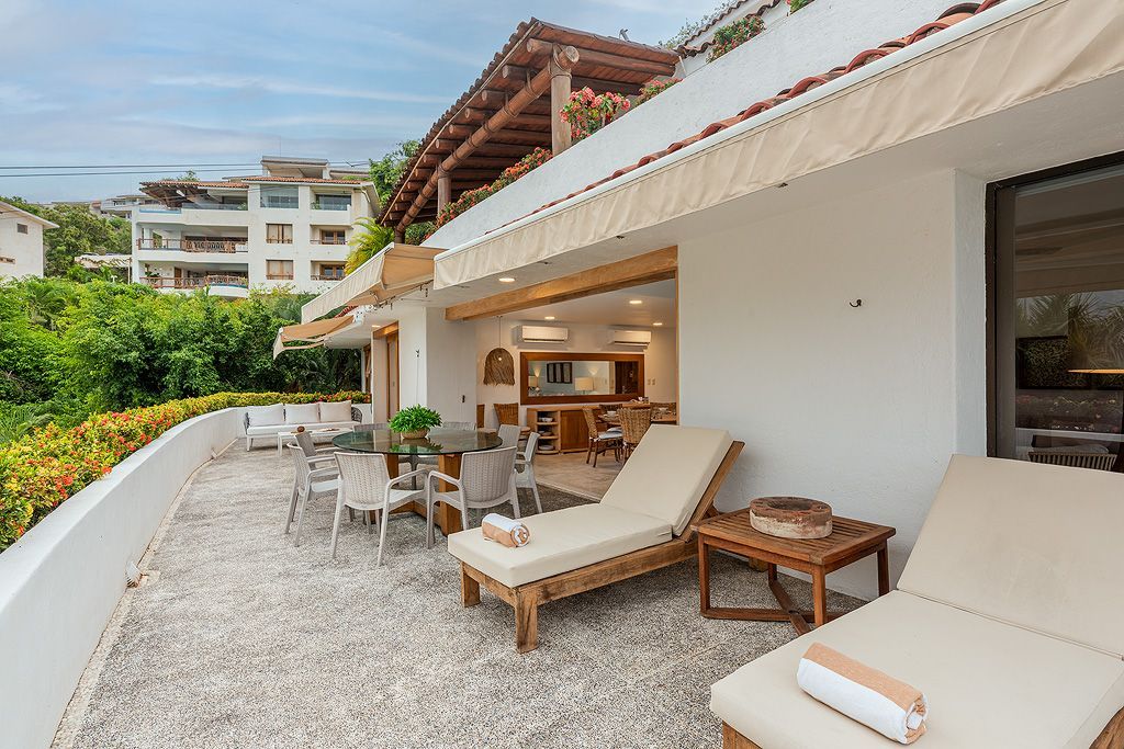 Terraza exterior con tumbonas, mesa y sillas, y una moderna villa blanca con patio abierto.