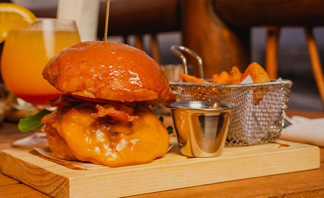 Hamburguesa y patatas fritas en una tabla de madera con salsa para mojar y bebidas al fondo.