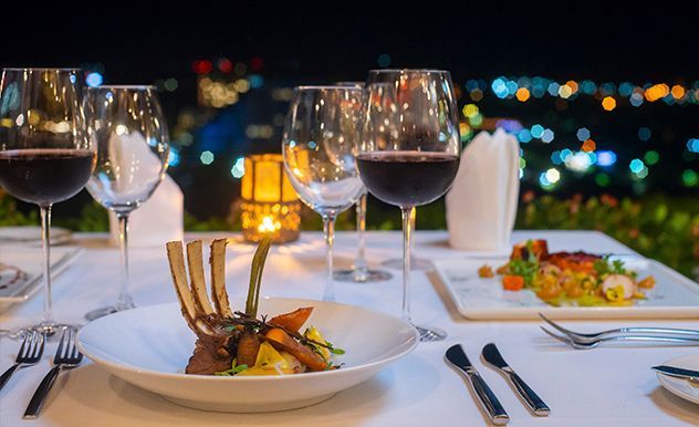Elegante mesa para cena a la luz de las velas con copas de vino y platos gourmet, con vistas a las luces de la ciudad por la noche.