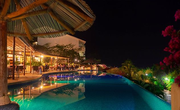 Piscina de lujo en un complejo turístico por la noche, con terraza comedor cálidamente iluminada y palmeras reflejadas en el agua azul.