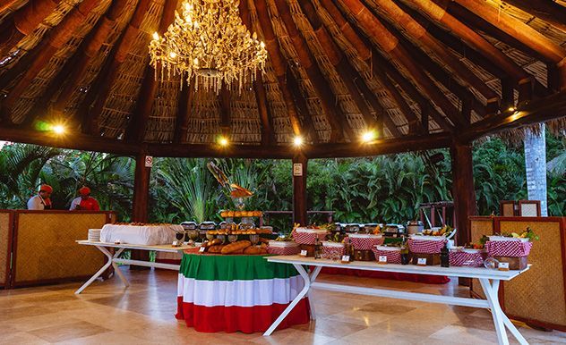 Banquete al aire libre con techo de paja, mesas blancas, flores y una lámpara de araña en un ambiente tropical.
