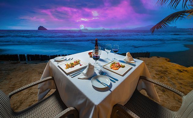 Cena romántica junto al mar al atardecer con vistas al océano, manteles blancos, velas y cielo púrpura.