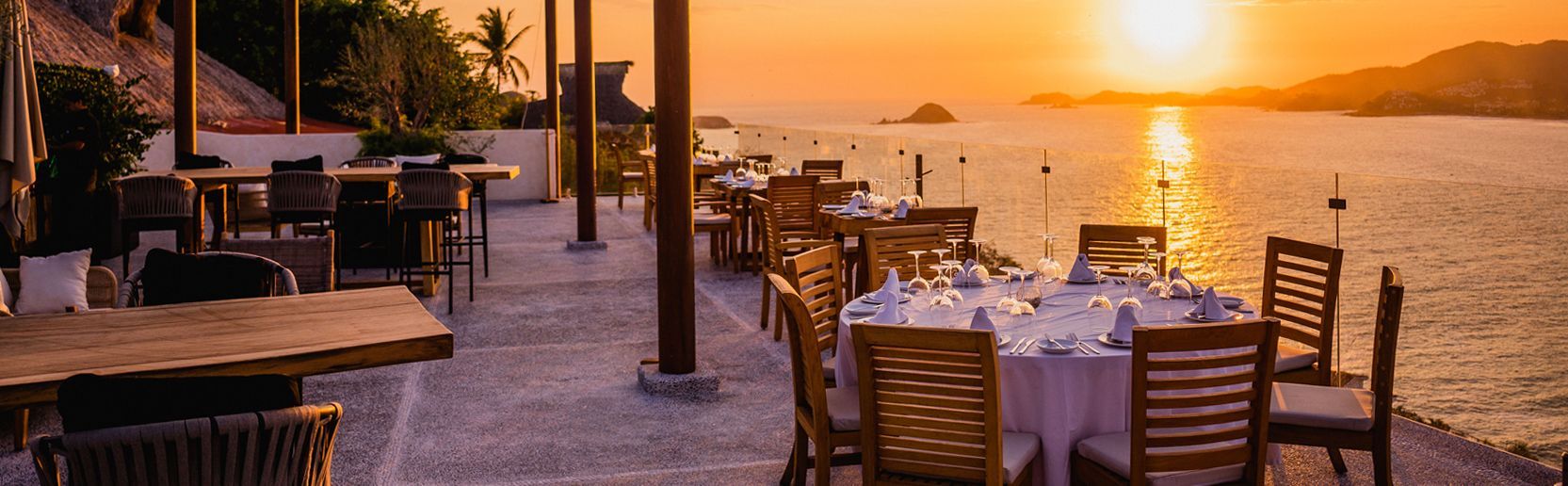 Terraza para cenar al atardecer junto al agua con manteles blancos, sillas de madera y una cálida luz dorada.