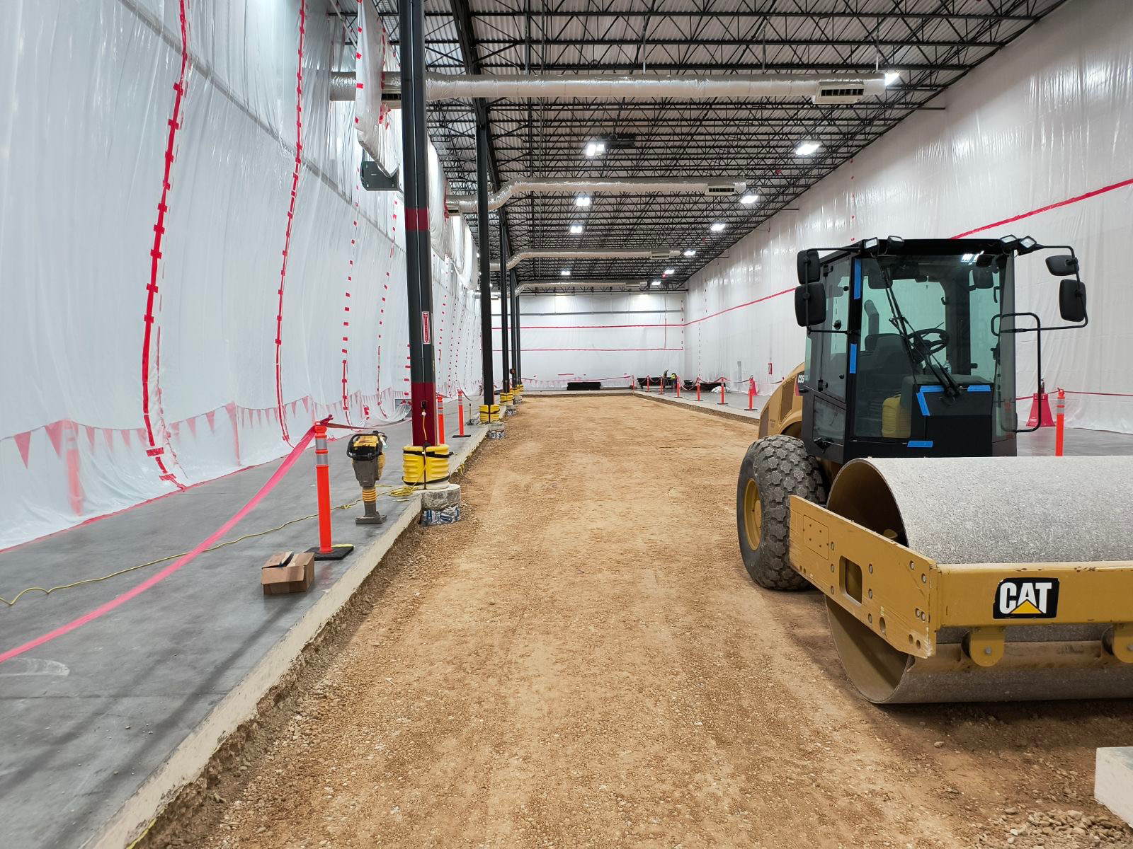 A cat roller is driving down a dirt road in a warehouse.