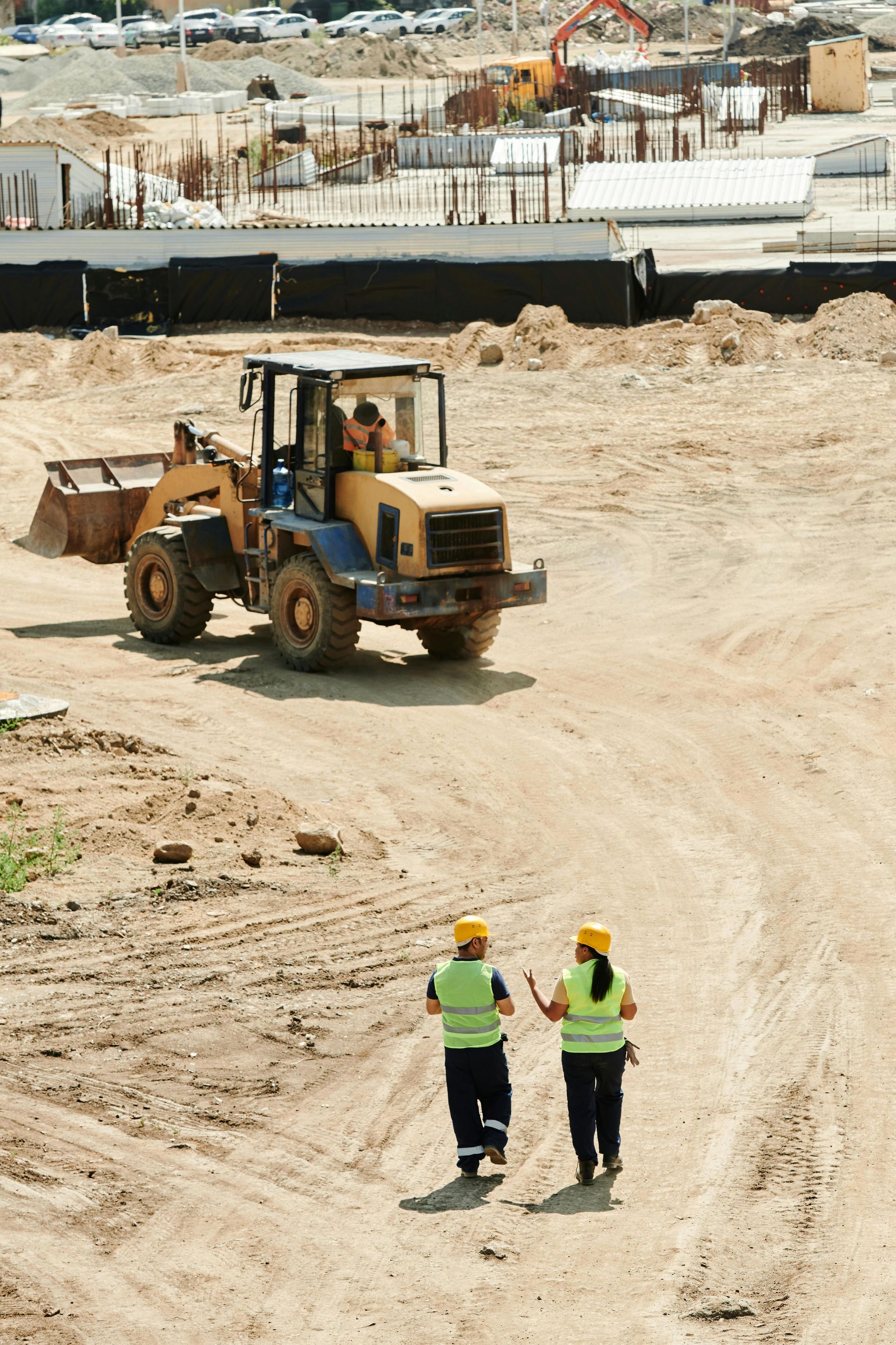 Two construction workers are walking in front of a bulldozer on a dirt road.