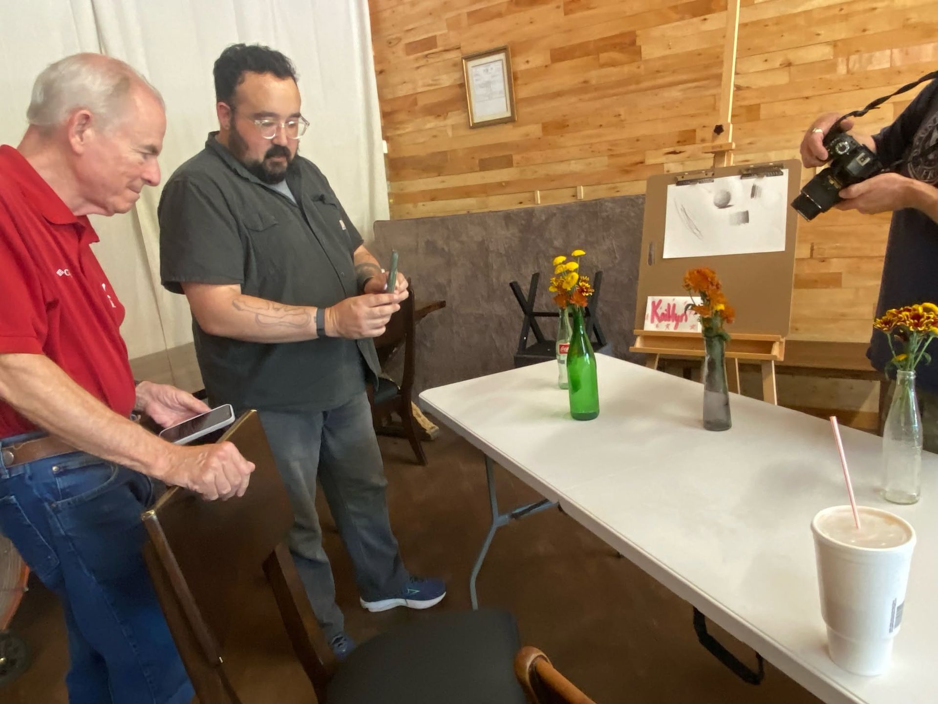 Three people at a table with flowers and drinks, looking at a display in a room with wood-paneled walls.