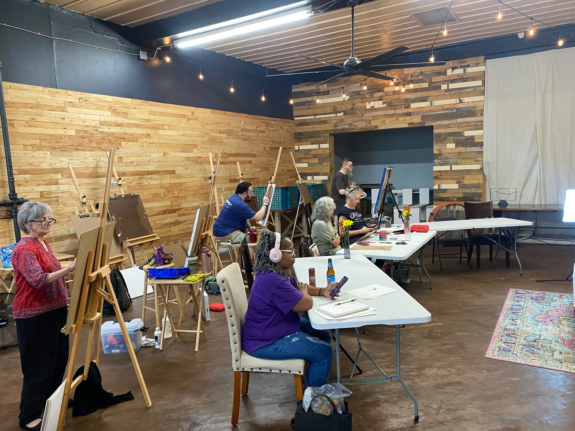 People painting at easels in a studio with wooden walls and string lights.