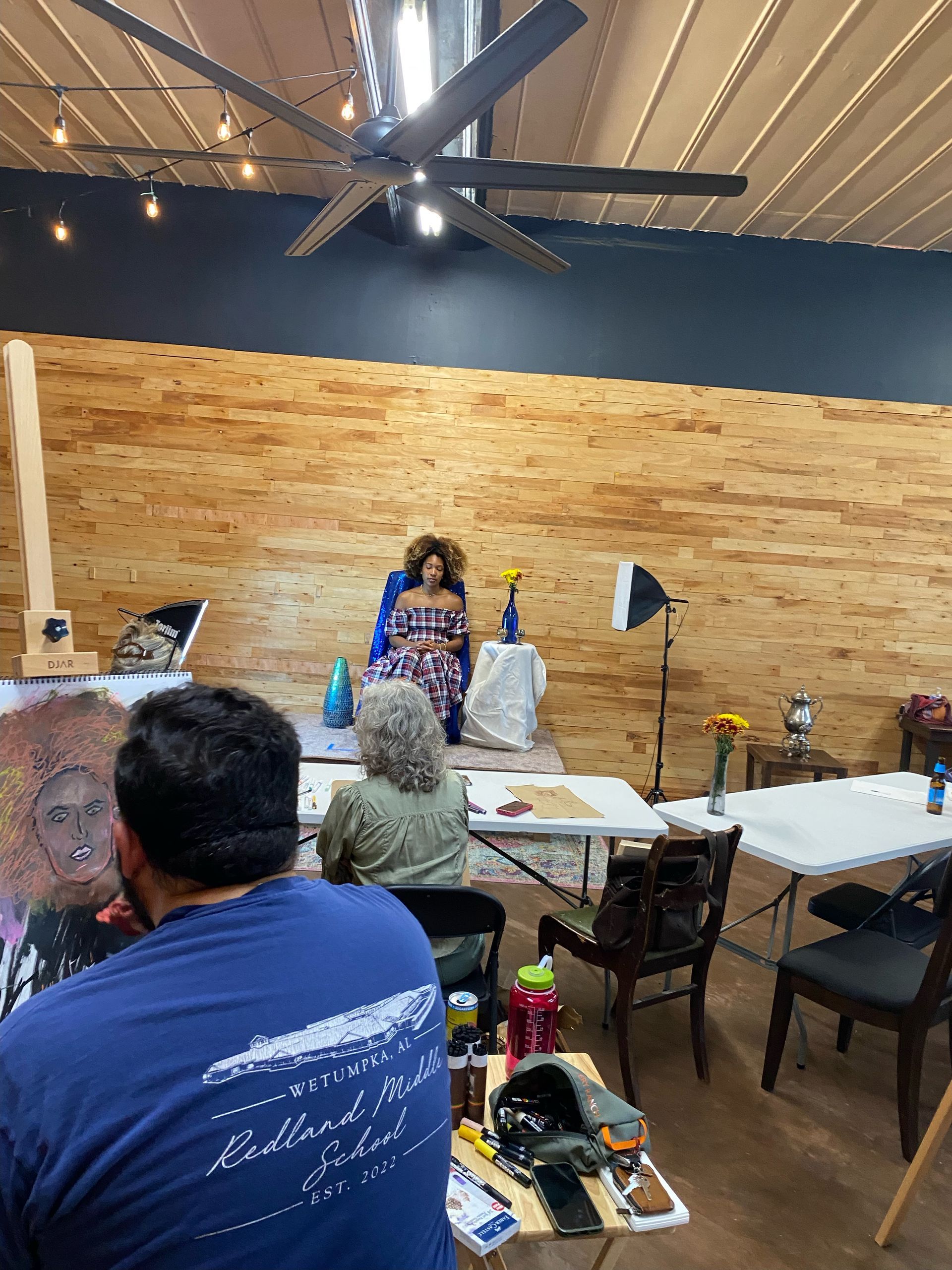 Speaker at front of rustic room, seated audience, exposed wood wall, ceiling fan, and tables with drinks.