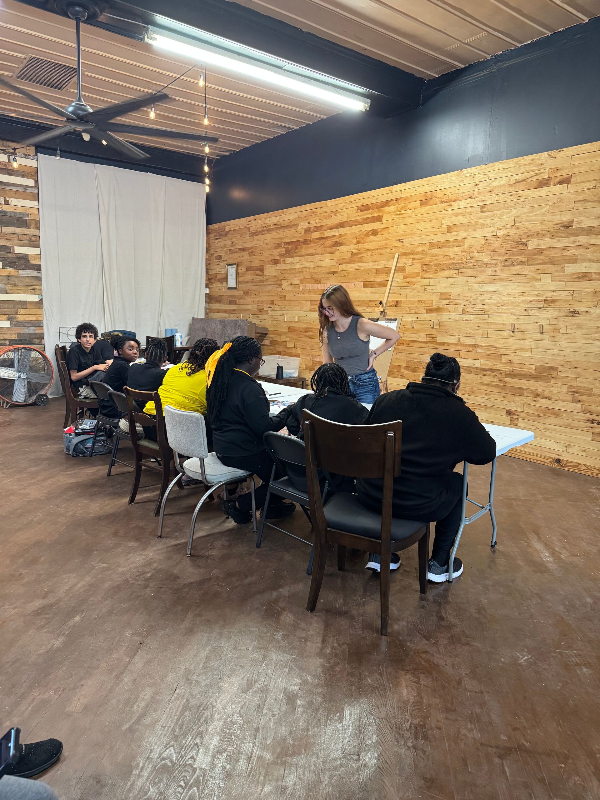 People seated at tables in a casual indoor meeting space with wood-paneled walls and a speaker at the front