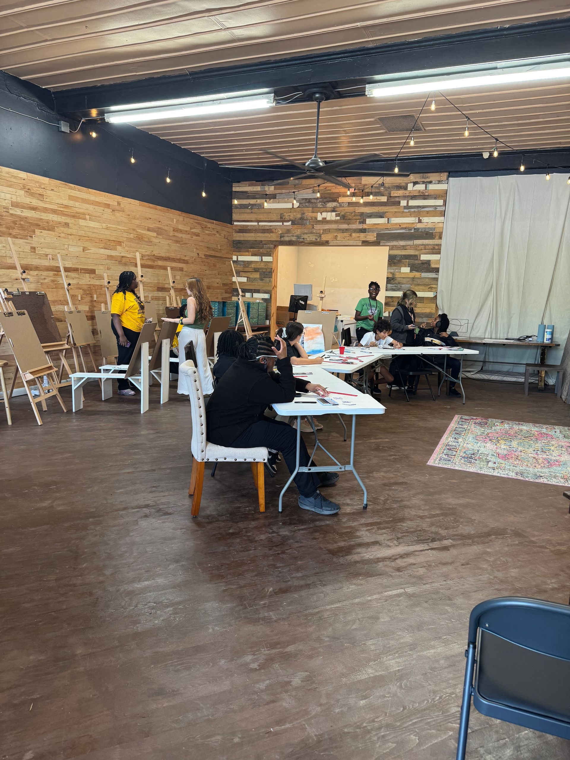 People seated at tables in a large hall with wood walls and a white drape, working or talking.