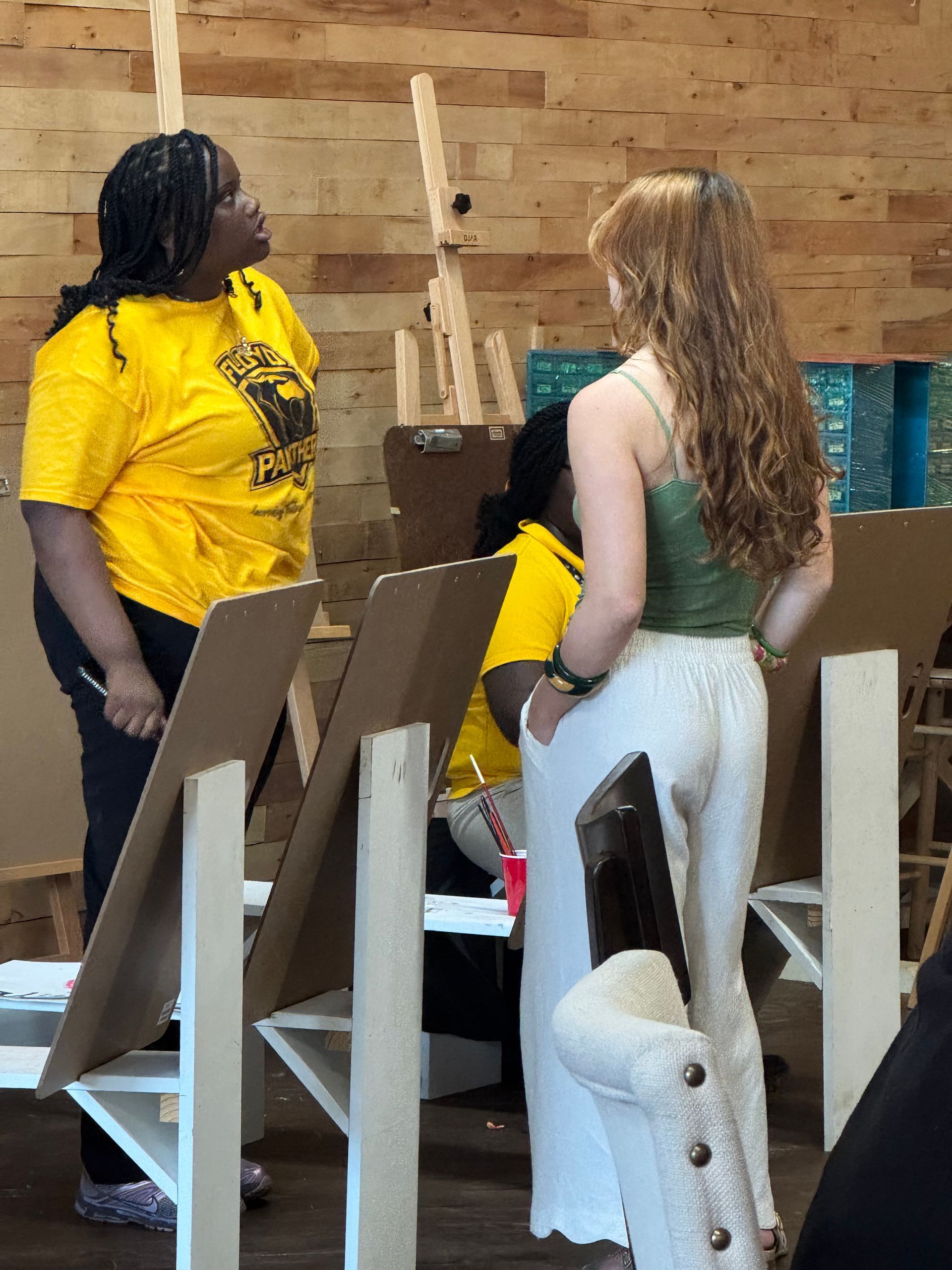 People standing among wooden panels in an indoor space, one in a yellow shirt and one in a light dress