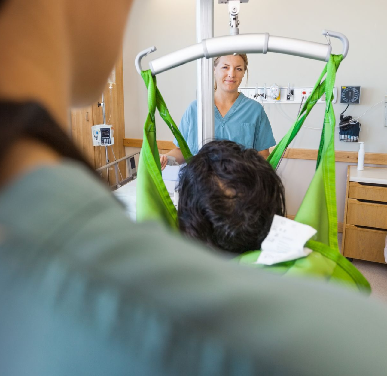 Patient suspended in a green sling as a nurse prepares a hospital lift transfer