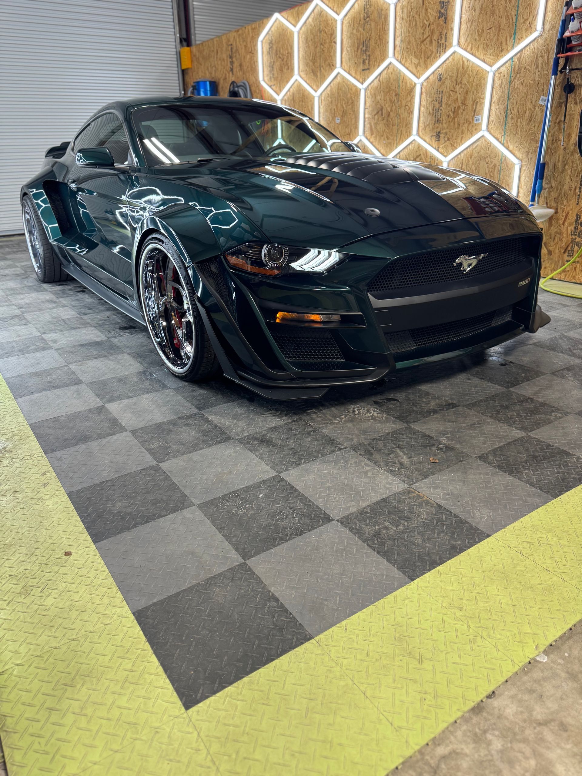 Dark green Ford Mustang, wide body kit, on checkered floor in garage.