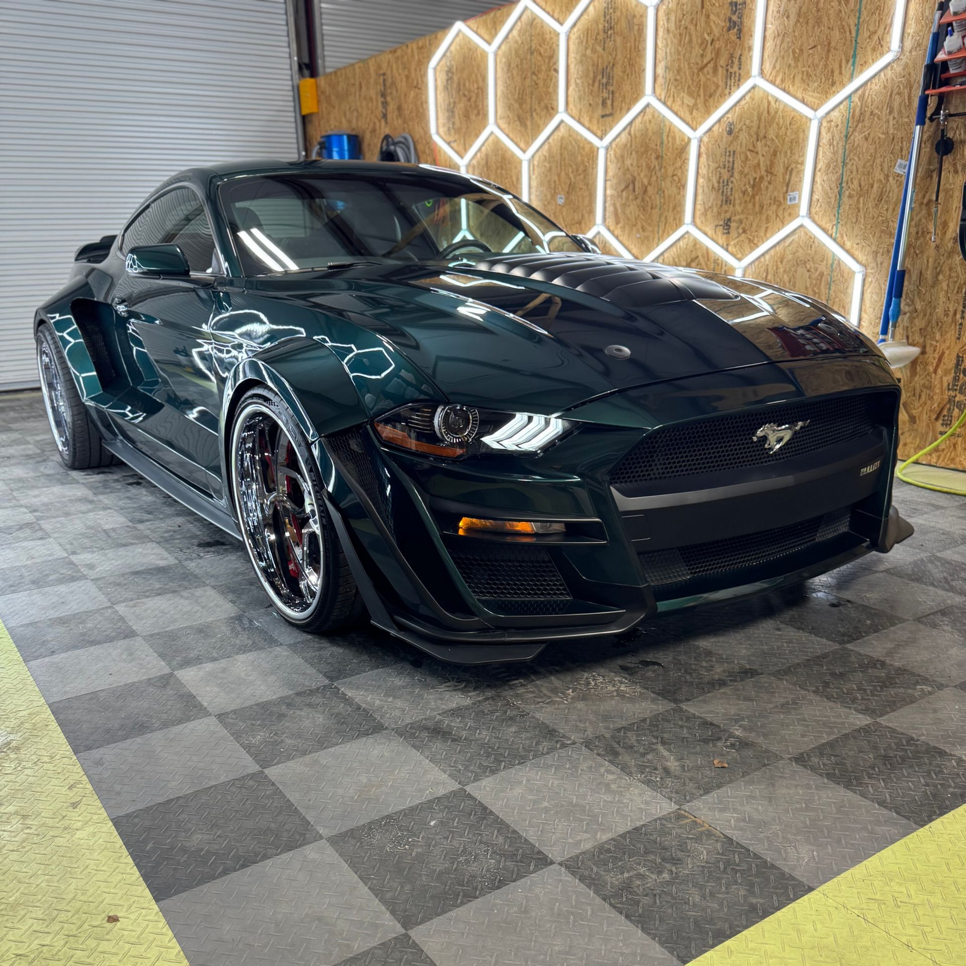 Dark green Ford Mustang, wide body kit, on checkered floor in garage.