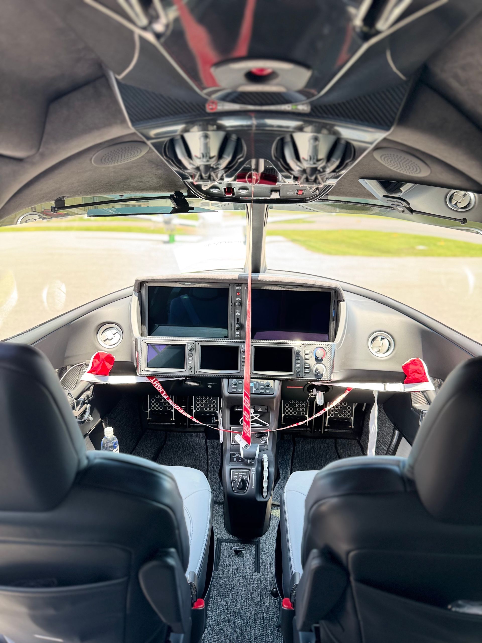 Interior of a race car cockpit with two seats, dashboard screens, and a roll cage visible through the rear window.