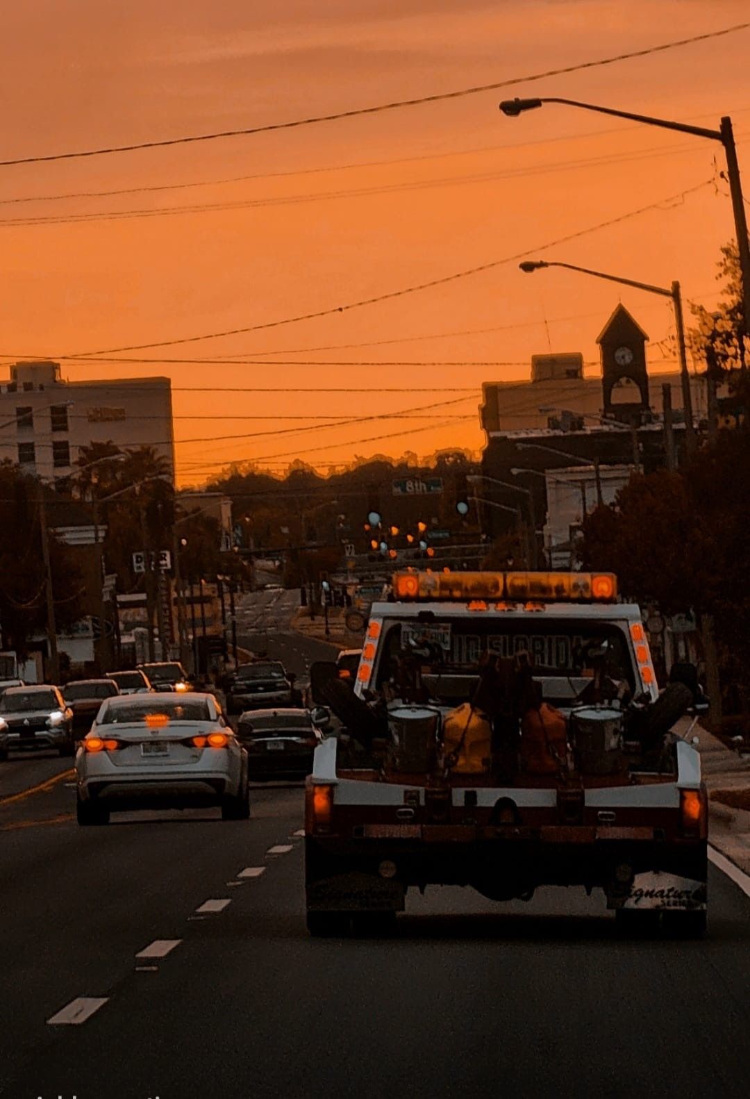 Tow truck driving on city street at sunset; orange sky.