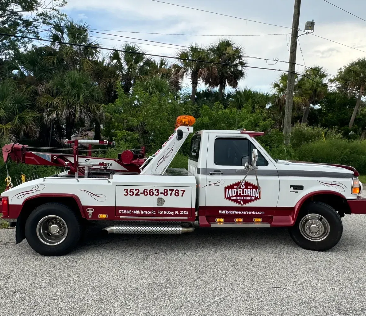 White and red tow truck parked on a gravel road with an orange light on top, trees in the background.