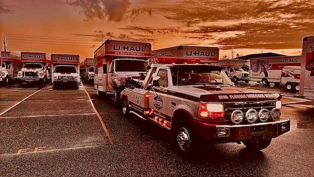 Tow truck hauling a U-Haul truck at sunset. Other U-Haul trucks are parked nearby.