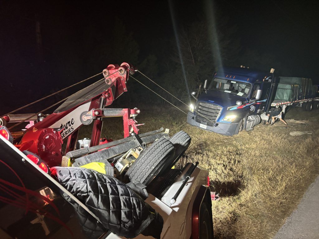 Tow truck pulling a semi-truck from a ditch at night. Red tow truck, dark blue semi, roadside setting.