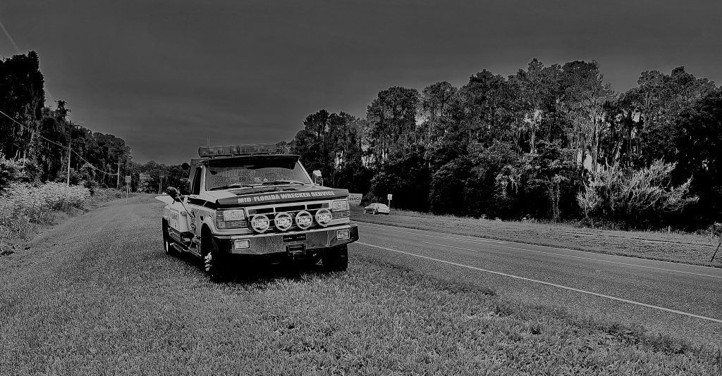Black and white photo of an old, rusted car parked in a field with trees in the background.