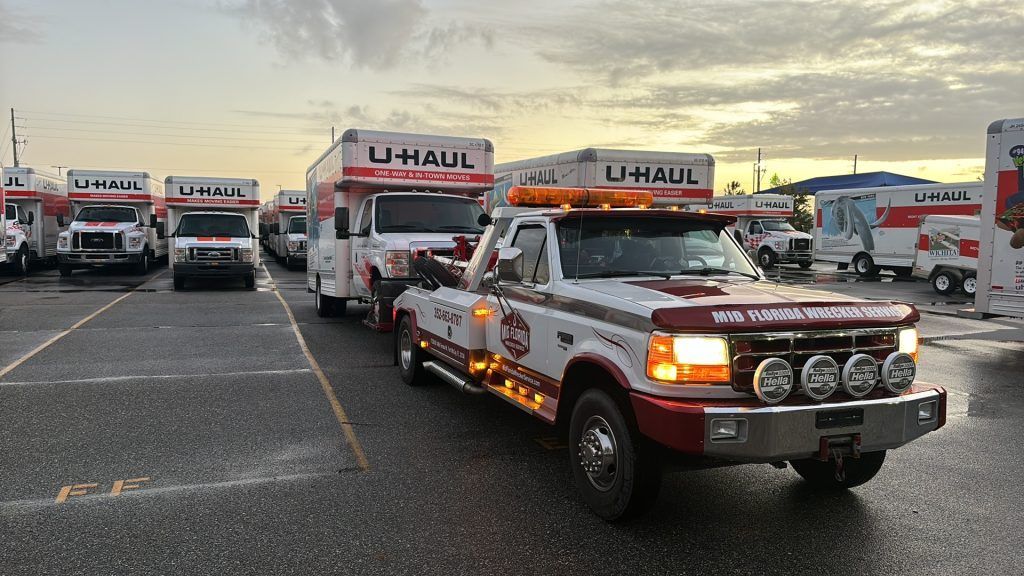 Tow truck towing a U-Haul truck. Other U-Haul trucks are parked in the background. Overcast sky.
