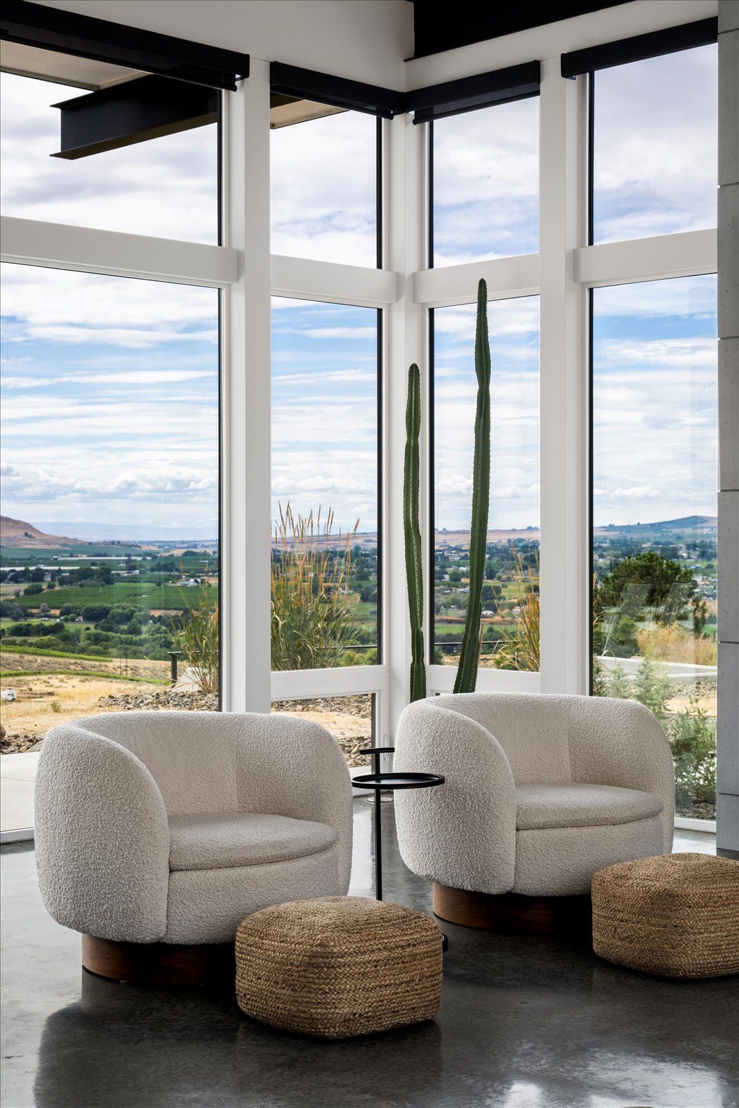 Two white, curved armchairs with woven footstools sit by a large window overlooking a valley.