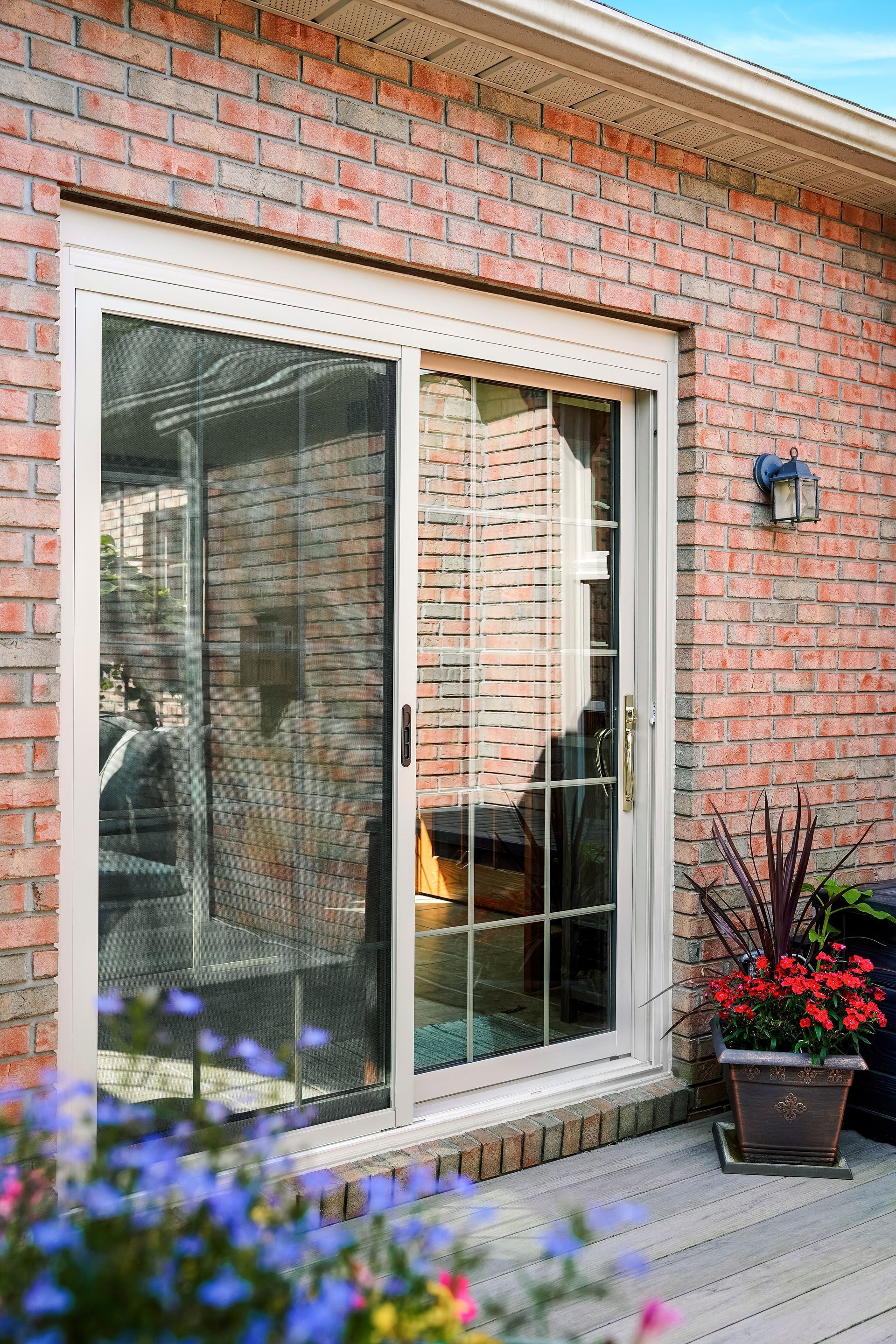 Brick patio door with a white frame beside a red brick wall, with a potted plant and flowers nearby.