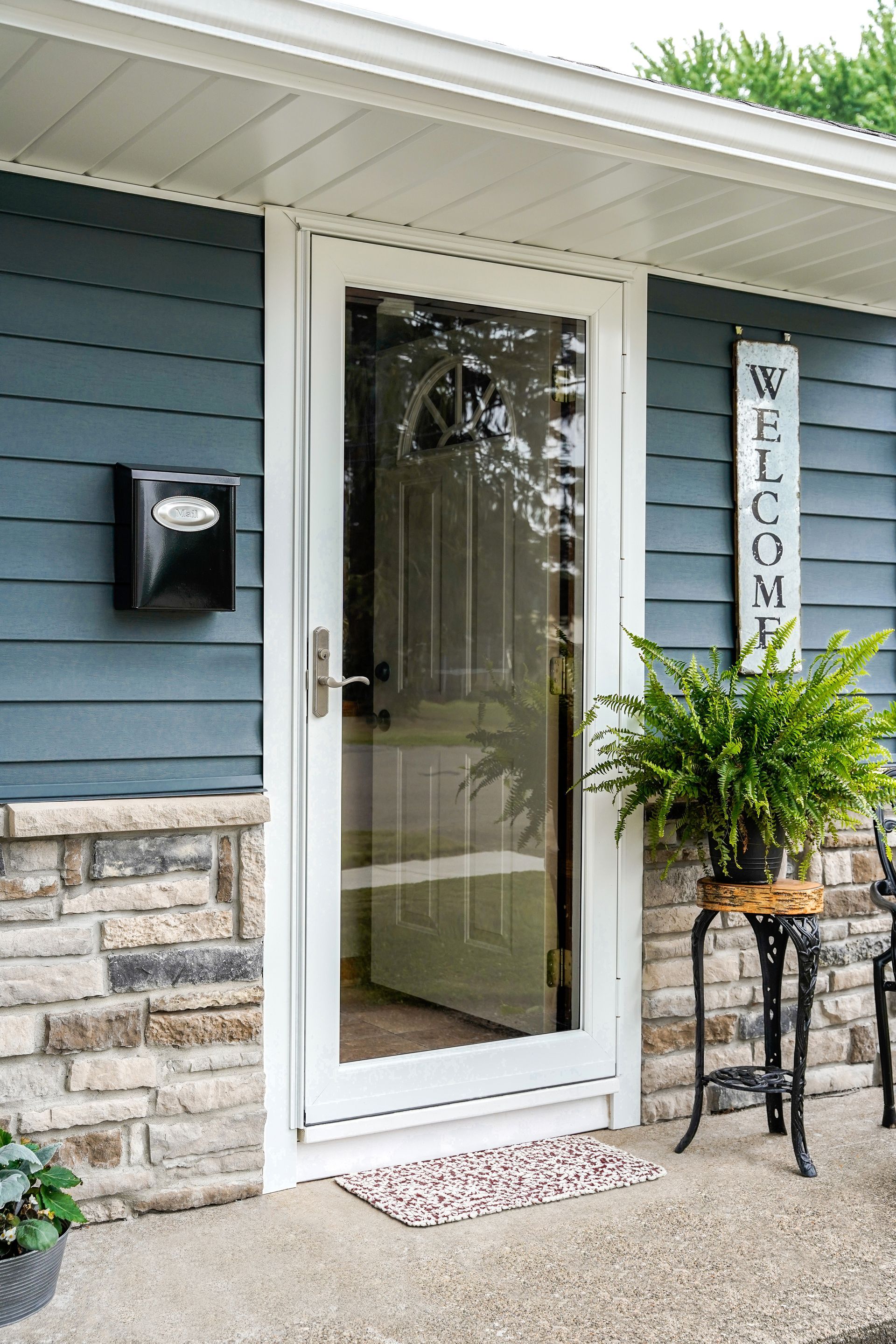 Front entry with glass storm door, welcome sign, and potted plant beside a blue-sided house