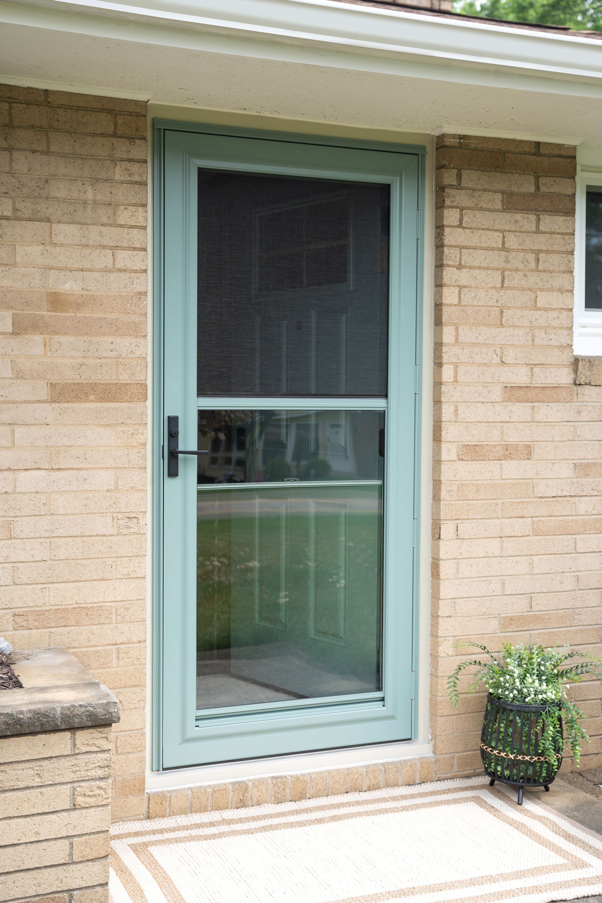 Teal screen door with glass panels on a tan brick house patio, beside a small potted plant.