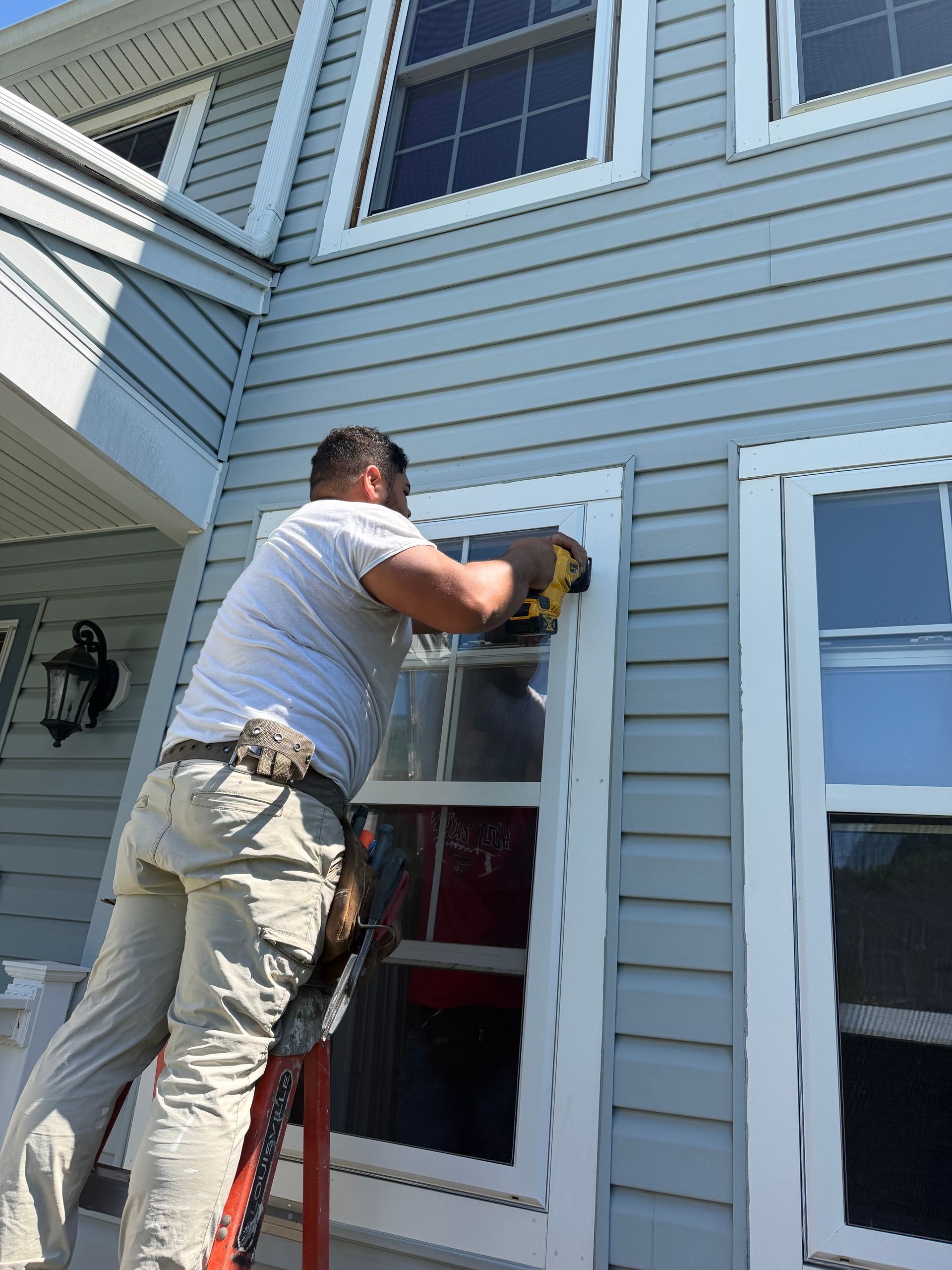 Person cleaning a second-story window on a gray house using a ladder and cloth