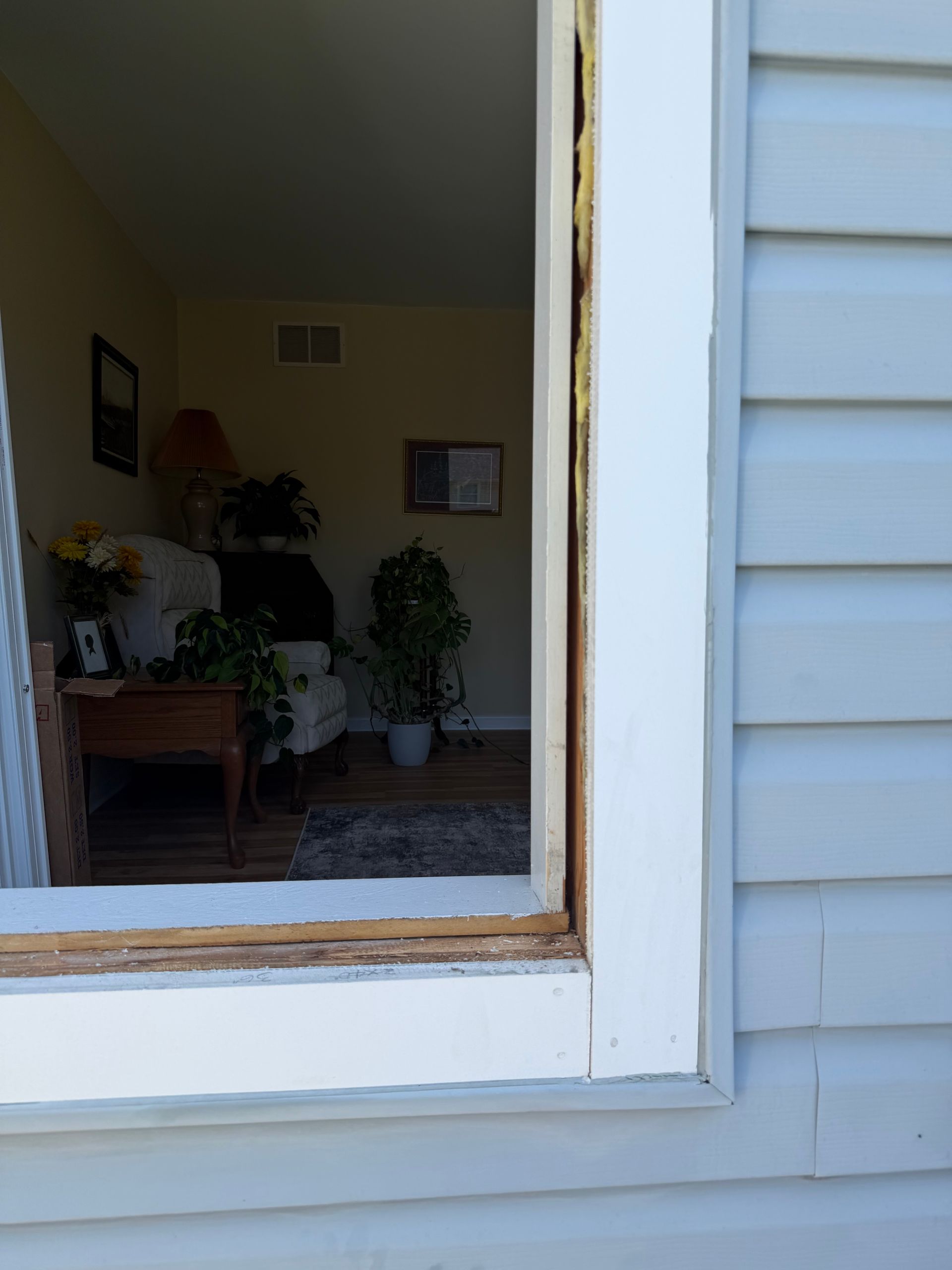 Open window beside white siding, looking into a softly lit living room with plants and framed wall art