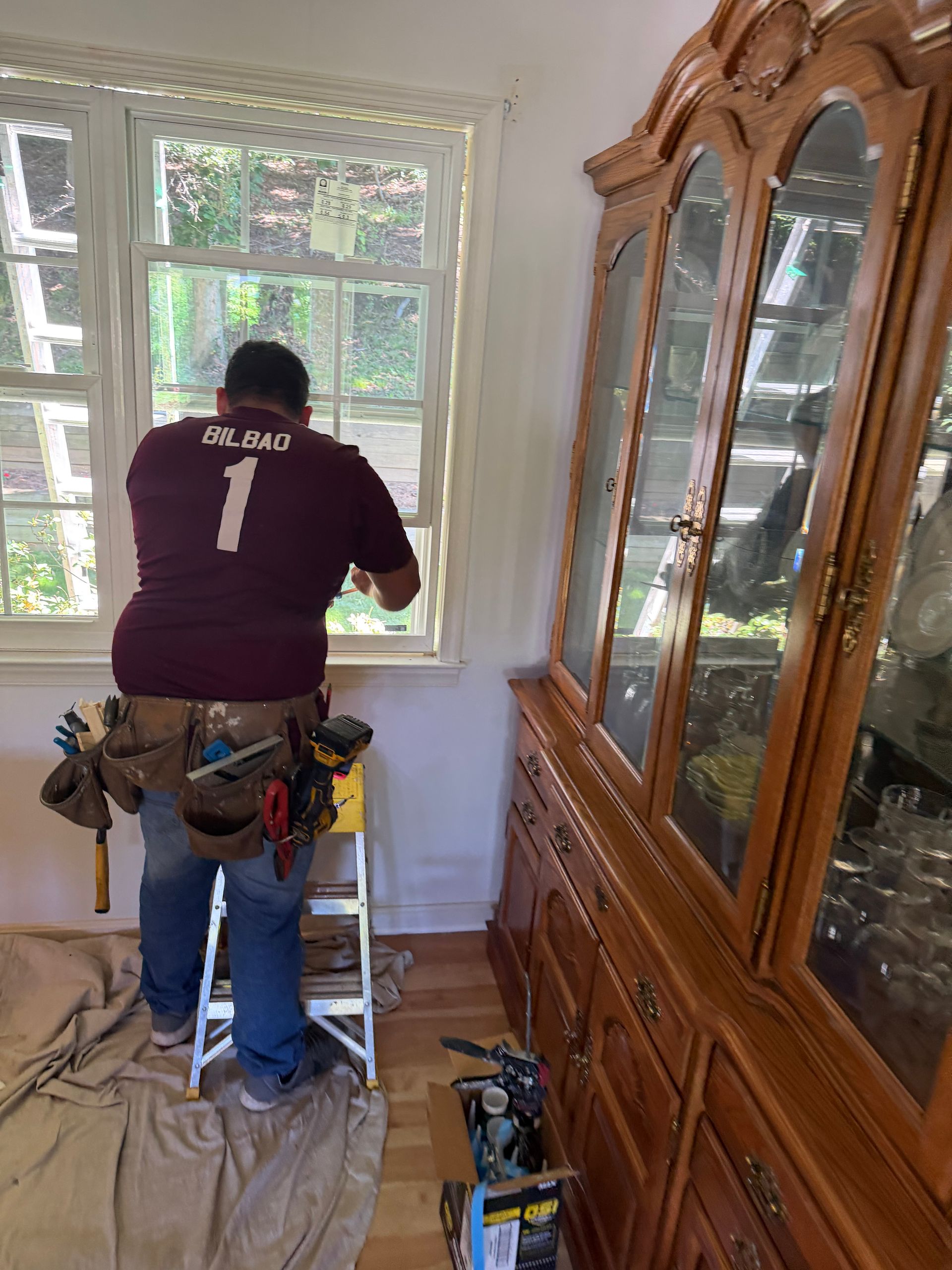 Person standing on a step ladder painting a window trim in a bright room, with tools on a belt.
