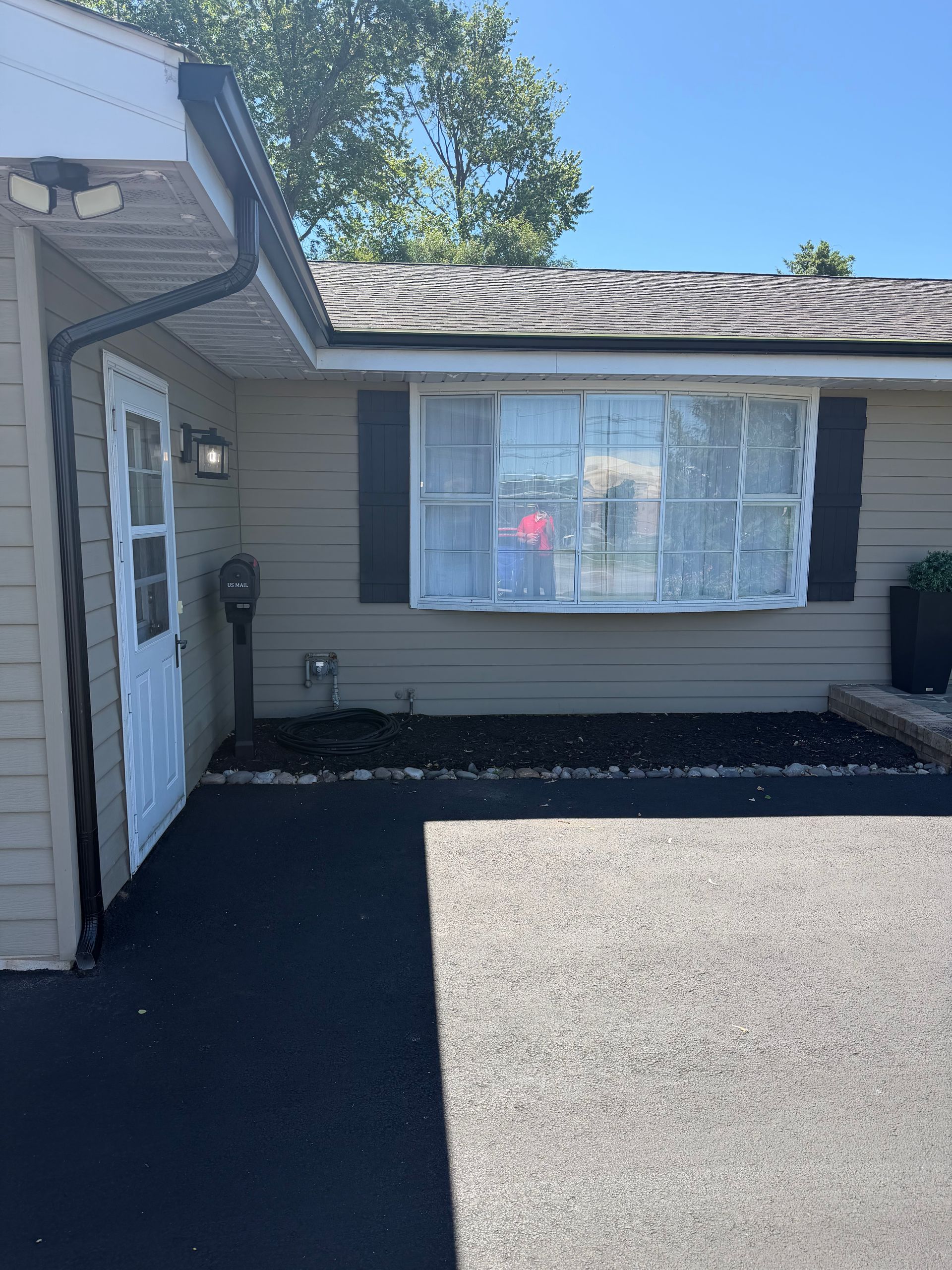Front of a beige house with a large window, dark shutters, and a paved driveway in sunlight
