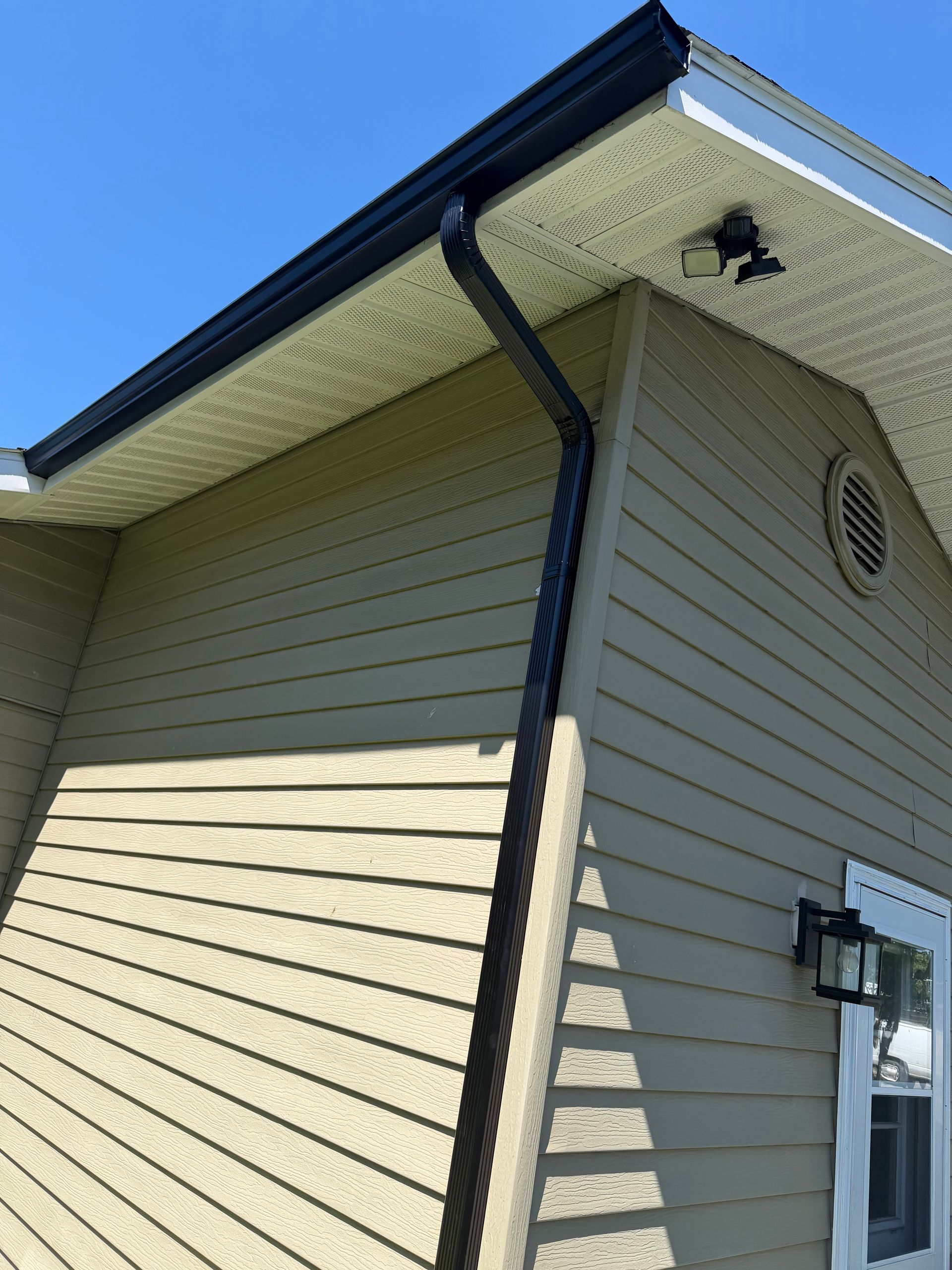 House exterior with beige siding, dark roof trim, and visible gutter downspout under blue sky