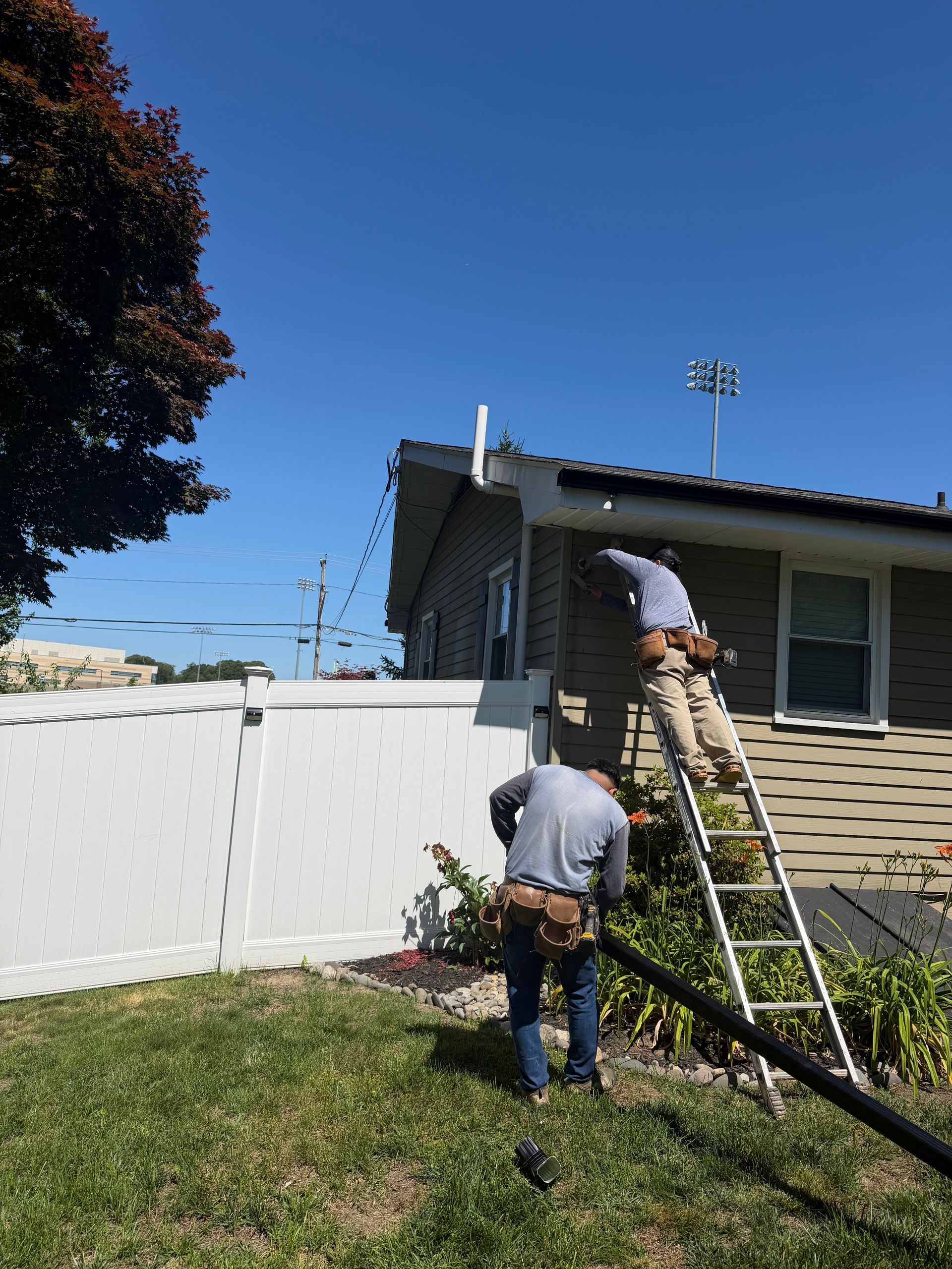 Two workers on ladders cleaning a house roof beside a white fence on a sunny day